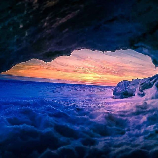 Dramatic sunset paints the sky seen from a cave opening over a sea of clouds. A surreal and tranquil landscape.