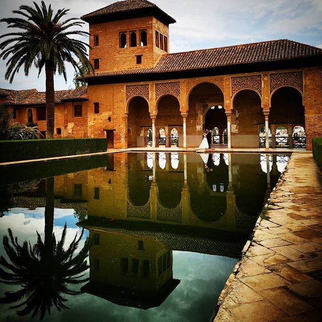 A bride and groom stand embraced in the Alhambra's courtyard, reflected in the pool, exuding romance and timeless elegance.