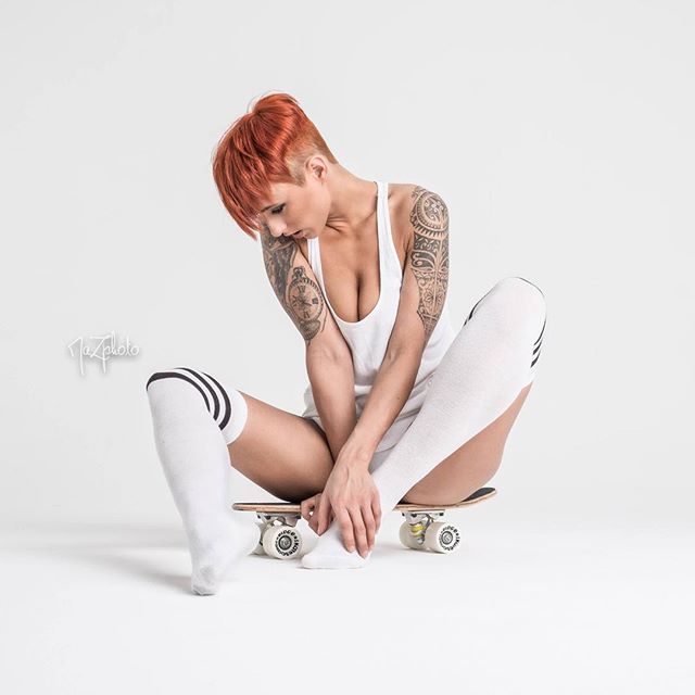 A tattooed woman in knee socks sits on a skateboard in a studio setting.