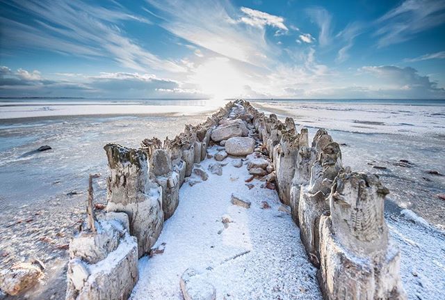 Winter seascape featuring wooden posts on a snow-covered beach under a bright sky.