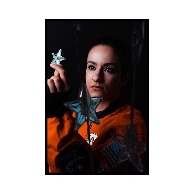 A woman in an astronaut jacket holds stars in a dreamy portrait against a dark background.