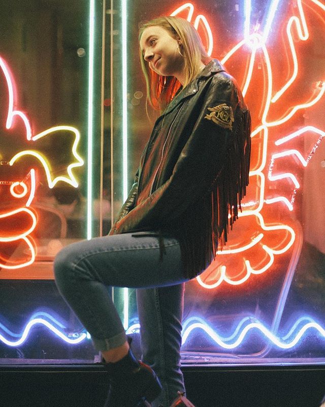 A stylish woman poses in a fringed leather jacket against a backdrop of vibrant neon signs.
