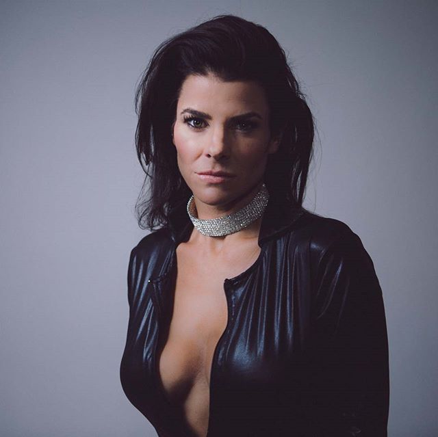 A woman in a black jacket and necklace poses confidently in a studio for a fashion or beauty editorial.