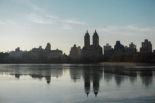 A serene cityscape reflects in the tranquil water of a lake, capturing the urban skyline under a clear, soft sky.
