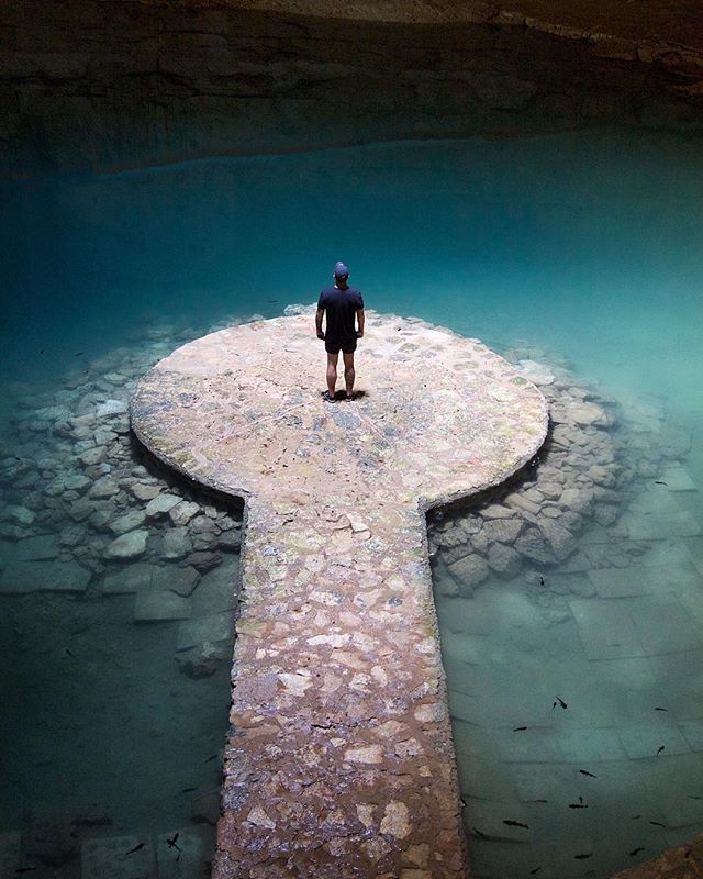 A man stands on a stone platform in a cenote, surrounded by clear blue water and rock formations.