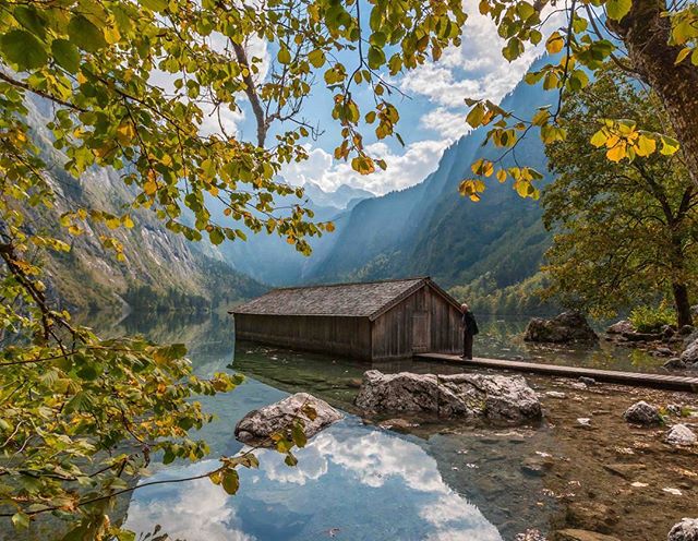 A boathouse sits on a tranquil lake surrounded by mountains and trees under a blue sky with clouds.