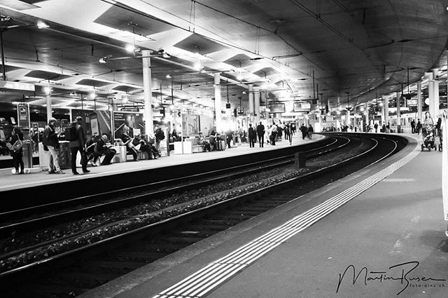 A black and white image of a train station platform with people waiting by the train tracks.