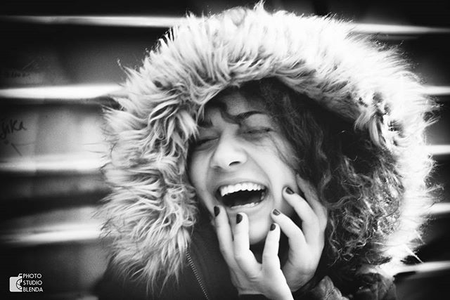 A cheerful woman with a fur hood laughs joyfully, her hands framing her face in this black and white portrait.