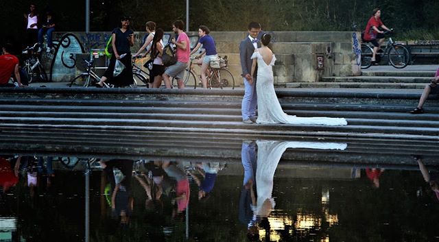 A wedding couple embraces on stairs near water, reflecting their image in a scenic urban park setting with many people around.