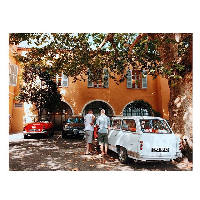 Two men admire classic cars parked on a tree-lined street in front of an orange building in Europe.