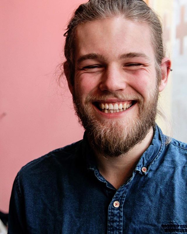 A happy man with a beard smiles in a medium shot wearing a denim shirt.