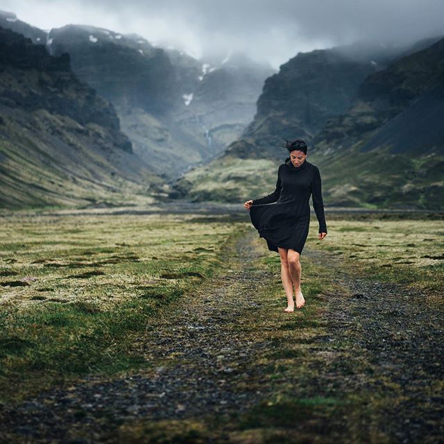 A woman in a black dress walks barefoot through a valley with mountains in the background.