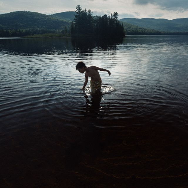 A young boy wades into a calm lake surrounded by mountains and trees on a cloudy day.