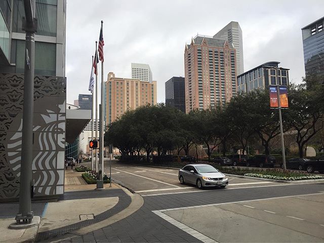 A silver car drives on a city street with buildings and flags in the background under a cloudy sky.