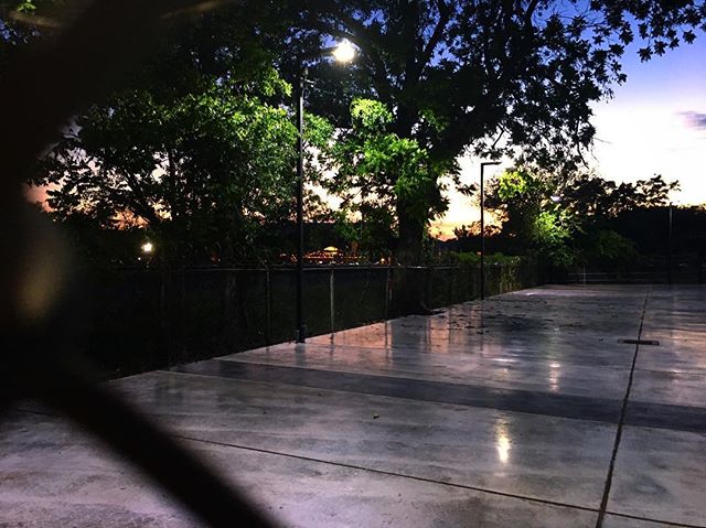 A quiet park scene at dusk with wet pavement, street lights, and trees under a fading sky.