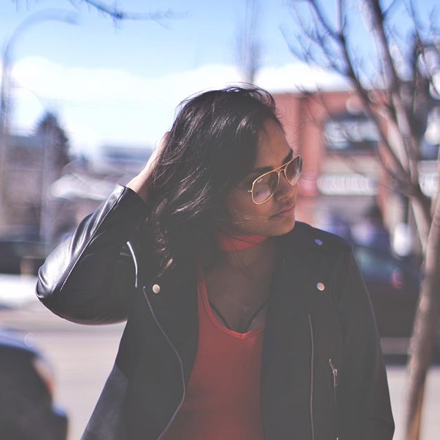 Woman with sunglasses and a leather jacket poses on a city street on a sunny day.