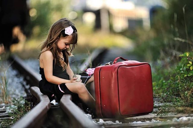 A young girl sits thoughtfully on a railroad track next to a vintage red suitcase, evoking a sense of travel and childhood memories.