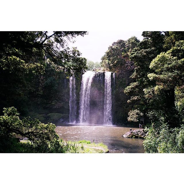 A scenic shot of a waterfall surrounded by lush trees and a small figure standing near the water.