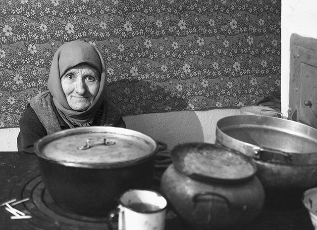 A woman in a headscarf sits near pots in a vintage kitchen, captured in black and white.