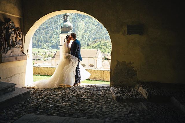A bride and groom share a kiss under a stone archway with a church in the background on their wedding day.