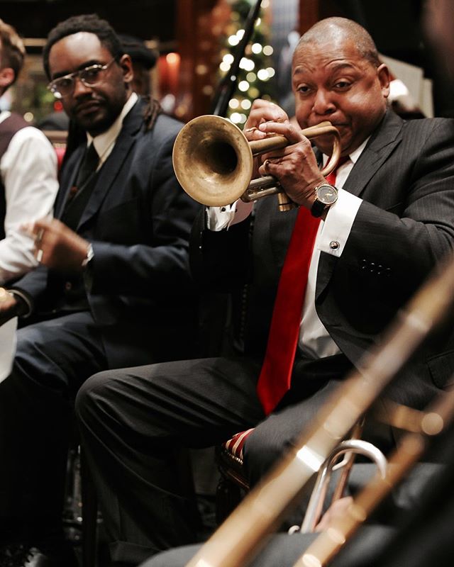 A man in a suit plays a trumpet during a jazz performance with an event backdrop.