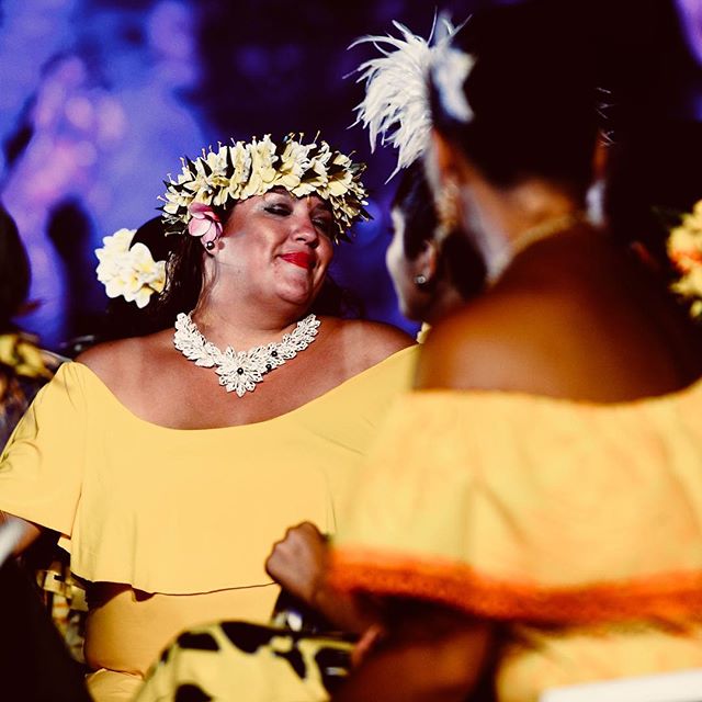 A smiling woman in a yellow dress and flower crown participates in a cultural performance.