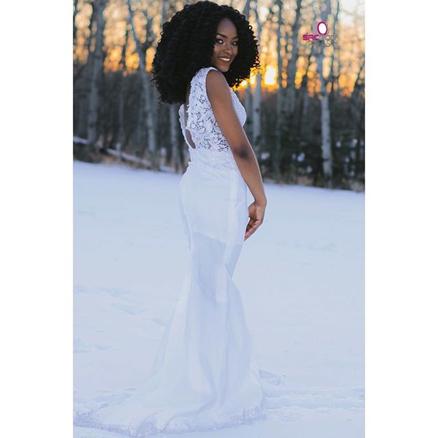 A woman in a white dress stands in a snowy forest at sunset, creating a serene and elegant scene.