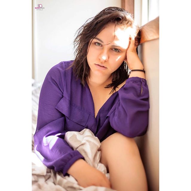 A brunette woman in a purple shirt sits by a window in a bright bedroom.