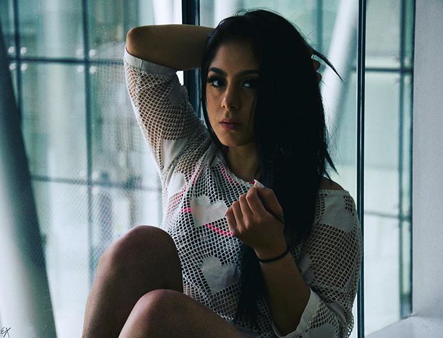 A young woman with dark hair poses by a window, wearing a white mesh top. 