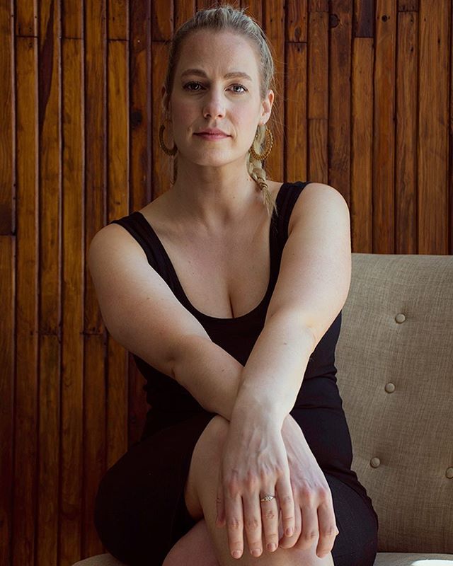 A blonde woman poses confidently in a natural light portrait with her arms crossed in a black dress.