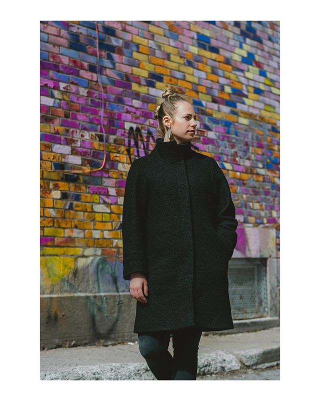 A woman in a black coat stands against a colorful brick wall on a city street.