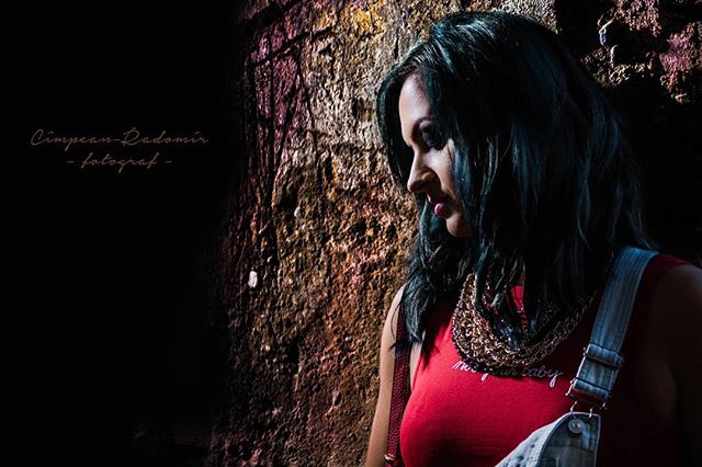 A brunette woman in a red top and overalls poses against a textured brick wall in soft, natural lighting.