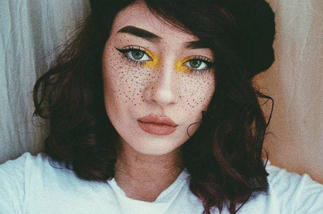 A brunette woman with freckles and soft makeup looks directly at the camera in a close-up shot.