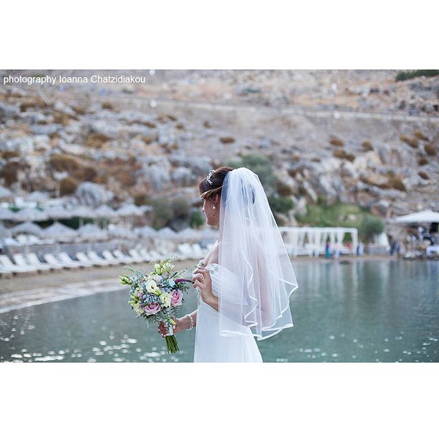 A bride in a white dress and veil holds a bouquet by the water on a beautiful coast.