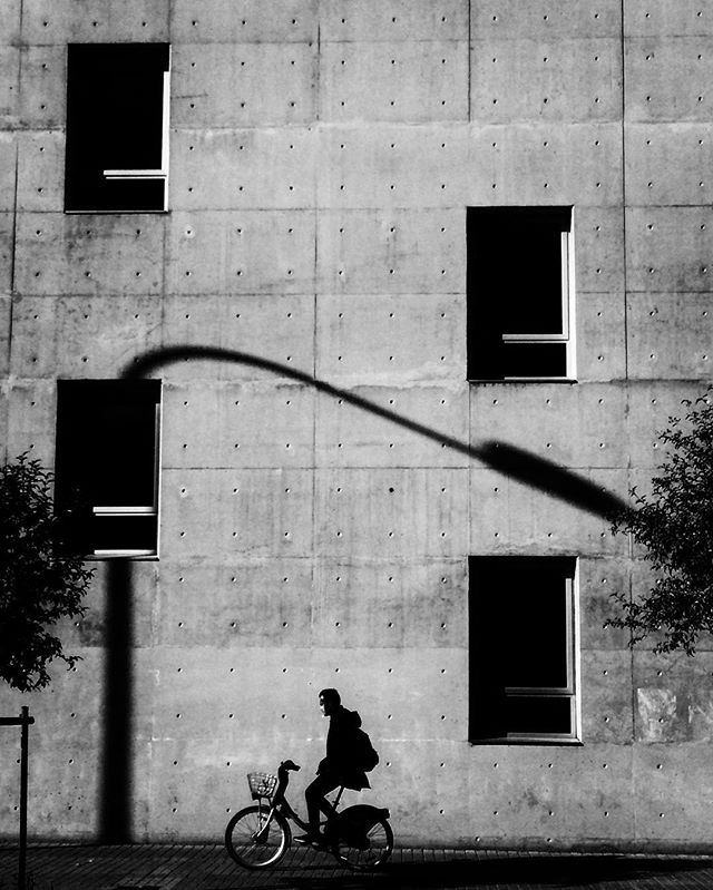 A cyclist rides in silhouette against a concrete building with geometric windows, creating a stark, minimalist urban scene.