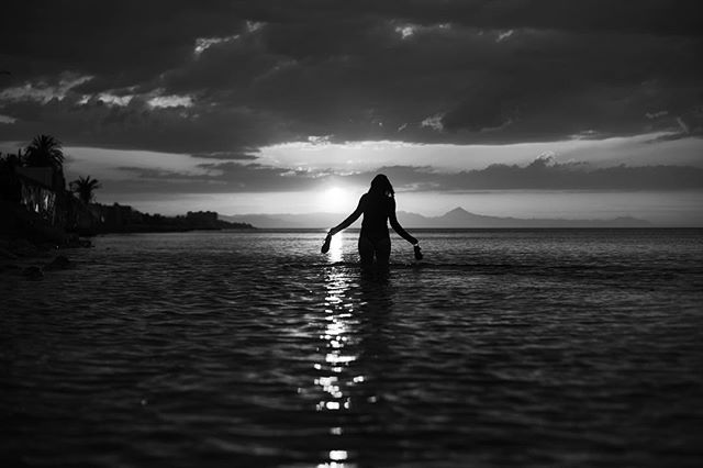 A woman walks into the ocean at sunset, a silhouette against the light and clouds.