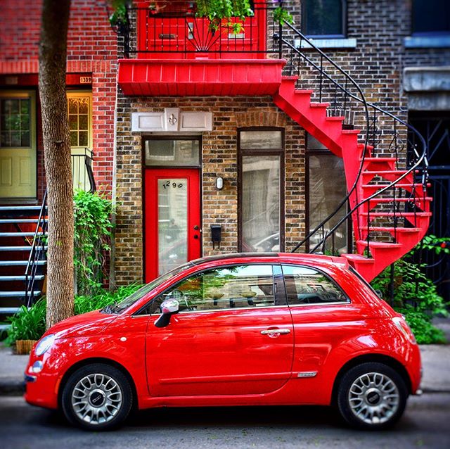 A red Fiat parked on a city street in front of a brick building with a red spiral staircase.