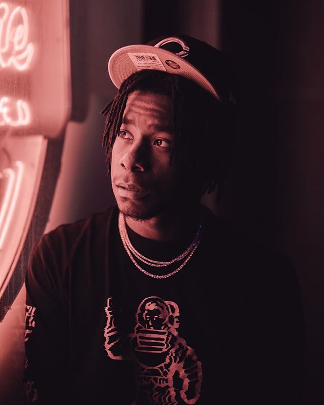 A stylish man poses in a dark studio, wearing a hat, chains, and a graphic tee. 