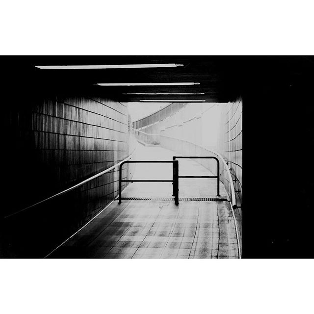 A black and white image of a walkway in a tunnel with railings leading towards a bright light.