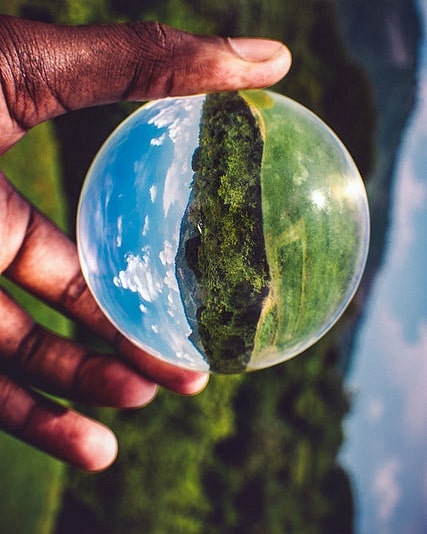 Hand holding a lensball reflecting a lush green landscape and blue sky, capturing the beauty of nature in a unique perspective.