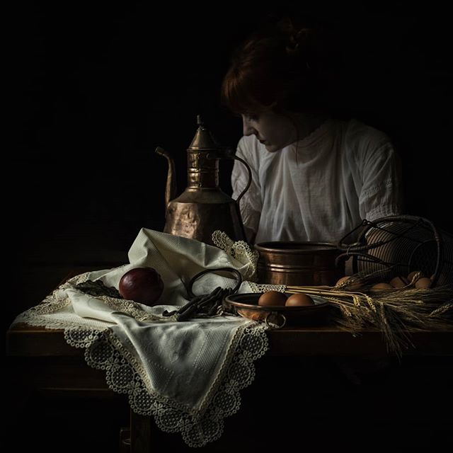 A woman is looking at a still life composition with a copper pitcher, an apple, and eggs on a lace-covered table.