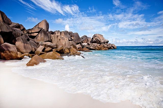 A scenic tropical beach with smooth rocks, clear turquoise ocean, and a blue sky with wispy clouds.