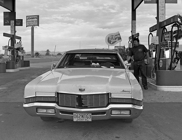 A vintage Mercury car is parked at a gas station while a man stands beside it in a black and white editorial image.