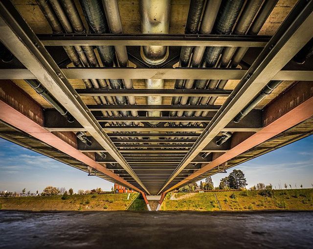 A low-angle shot showcases the intricate underbelly of a bridge with pipes and beams against a blue sky and green landscape.