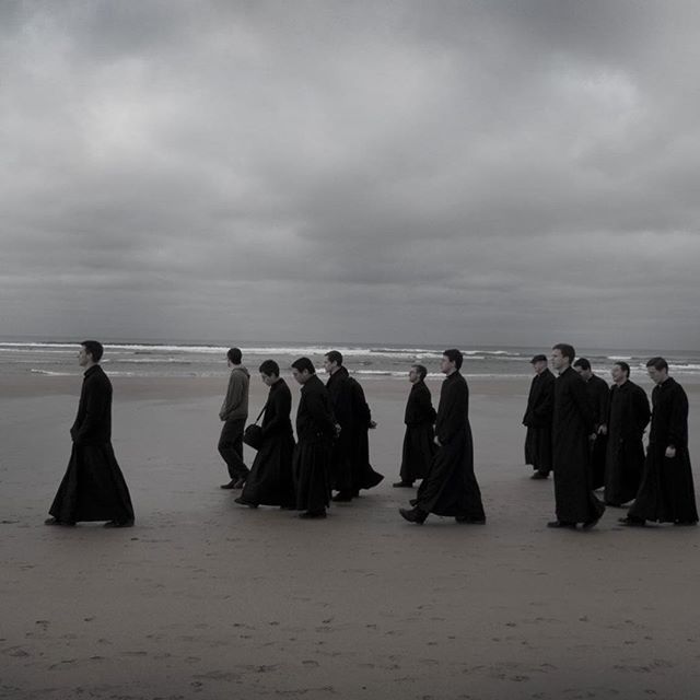 A group of clergy members walk along a beach under a cloudy sky in a monochrome image.