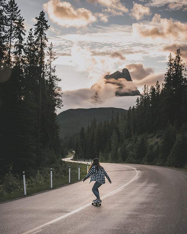 A woman skateboards down a winding road surrounded by trees and mountains under a cloudy sky.