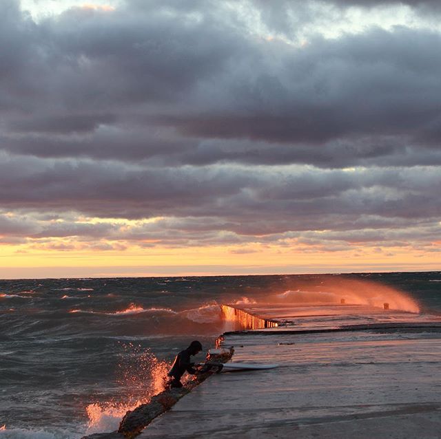 A person stands on a pier as waves crash at sunset, creating a moody and intense seascape.