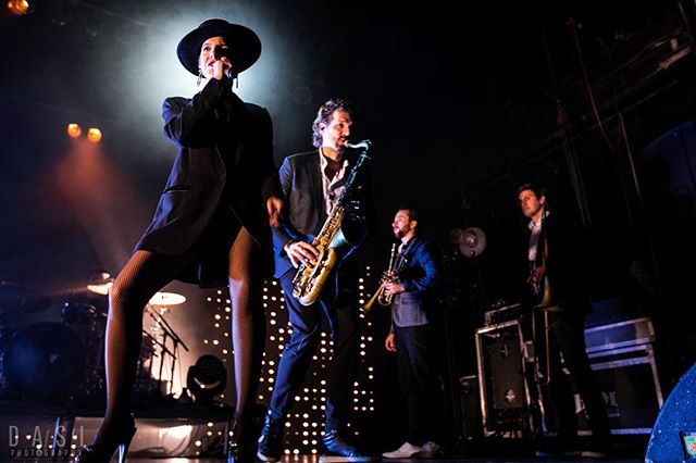 A woman sings on stage with a band playing instruments during a concert.