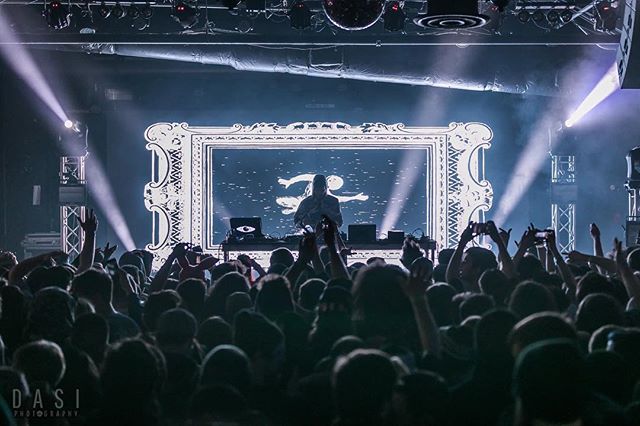 A lively concert featuring a DJ on stage before a large crowd, illuminated by dramatic stage lighting.
