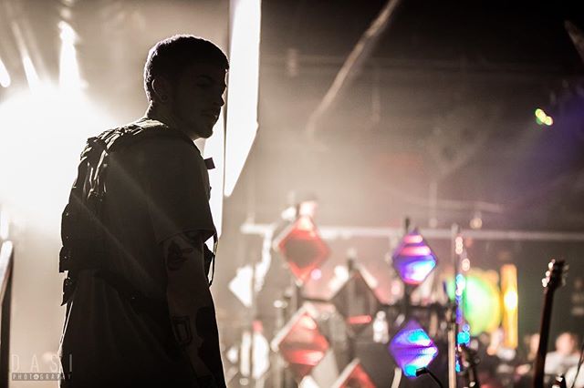 A silhouetted man stands on stage with a backpack, possibly before or after a music performance.